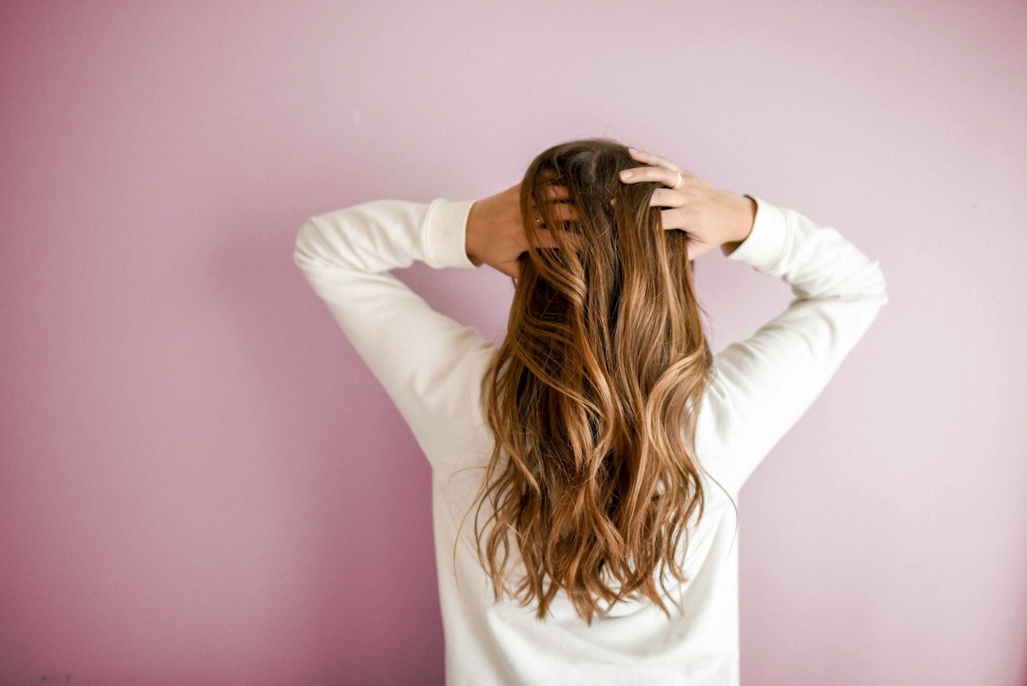 A woman in a white sweater running her hands through long, brightened brown hair against a pink wall